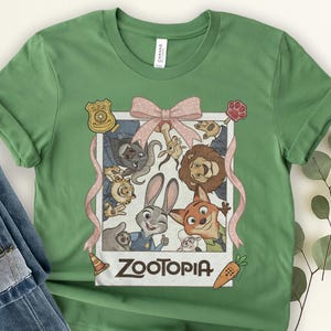 K&ouml;nnte beinhalten: Gr&uuml;nes T-Shirt mit einer Grafik von Zootopia-Charakteren in einem Polaroid-Rahmen und einer rosa Schleife. Das Zootopia-Logo befindet sich unten. Das T-Shirt besteht aus einem weichen Material.