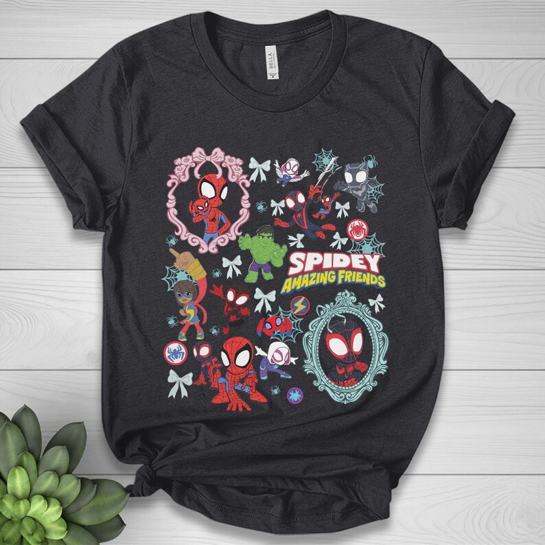 May include: Dark gray t-shirt featuring a colorful graphic of Spider-Man and other characters from "Spidey and His Amazing Friends." The design includes the show's title and various illustrations of the characters within decorative frames and bows.