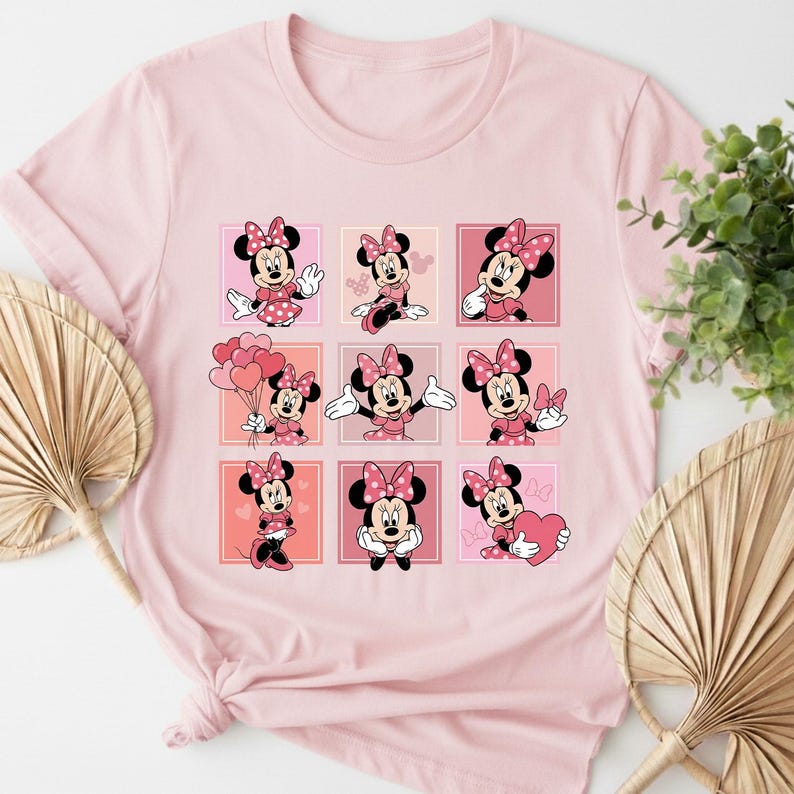 May include: Pale pink t-shirt with a grid of nine Minnie Mouse illustrations. Each Minnie Mouse is depicted in a different pose, wearing a pink dress and bow. The shirt is made of a soft material.