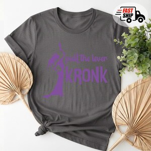 May include: Dark gray t-shirt with a purple graphic of a character and the text "pull the lever KRONK". The shirt is folded and knotted at the bottom. A Fast Ship logo is in the upper right corner.