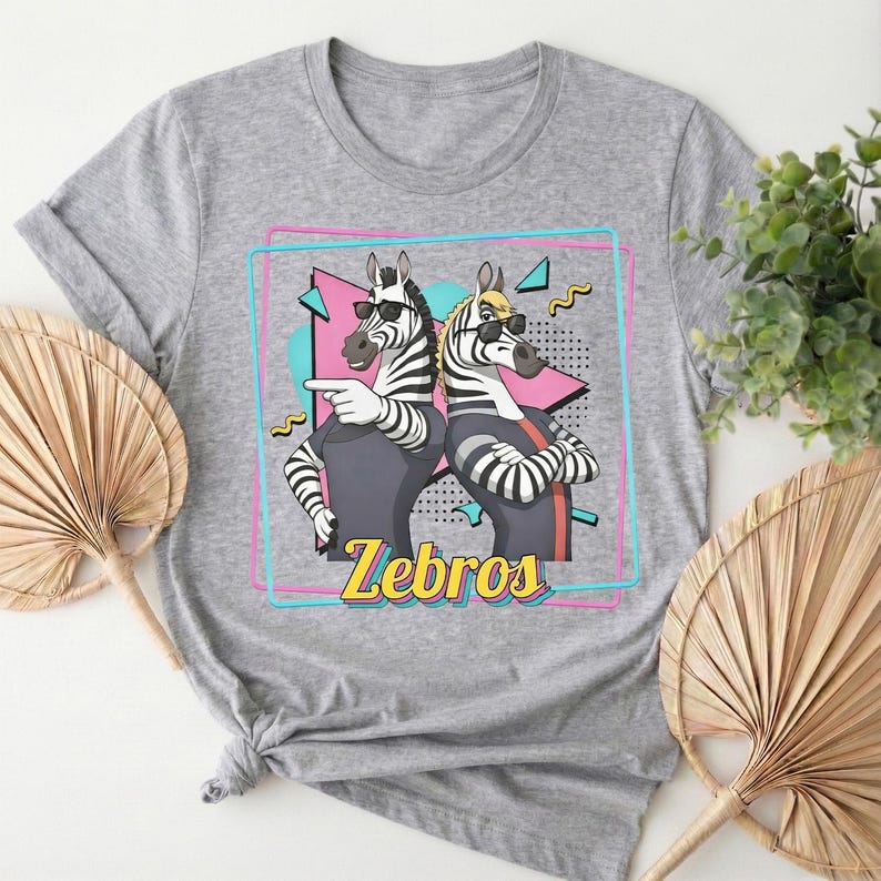 May include: A gray t-shirt featuring a graphic of two zebras wearing sunglasses and posing. The zebras are inside a pink and blue outlined square with the word "Zebros" in yellow. The shirt has short sleeves.