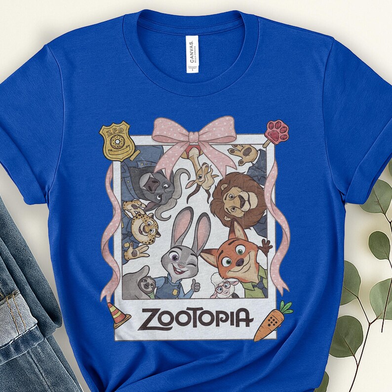 K&ouml;nnte beinhalten: Ein blaues T-Shirt mit einem Aufdruck von Charakteren aus dem Animationsfilm Zootopia. Das Design umfasst eine rosa Schleife, ein Polizeiausweis, einen Pfotenabdruck und eine Karotte. Das Wort "Zootopia" ist unten im Design wei&szlig; gedruckt.