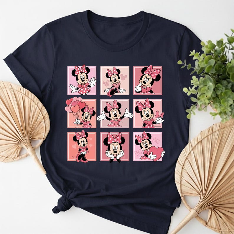 May include: Dark navy t-shirt with a grid pattern of Minnie Mouse illustrations. Each square displays Minnie in a pink dress and bow, set against a pink backdrop. The design incorporates hearts and balloons, offering a playful and cheerful appearance.