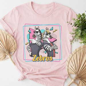 May include: Pink t-shirt featuring a graphic of two zebras wearing sunglasses, with the word "Zebros" in yellow. The zebras are in a retro-style design with a teal and pink border.