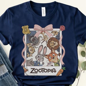 K&ouml;nnte beinhalten: Marineblaues T-Shirt mit einer Grafik von Zootopia-Charakteren in einem Polaroid-Rahmen und einer rosa Schleife. Das Wort "Zootopia" ist unten aufgedruckt. Das T-Shirt ist aus weichem Material.