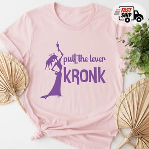 May include: A light pink t-shirt featuring a purple graphic of a character with the text "pull the lever KRONK". The shirt has a crew neck and short sleeves. The design is centered on the front of the shirt.