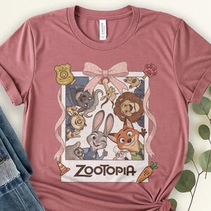 K&ouml;nnte beinhalten: Staubrosa T-Shirt mit einer gerahmten Grafik von Zootopia-Charakteren, darunter Judy Hopps und Nick Wilde. Die Grafik ist mit einer rosa Schleife und einem Band verziert. Das Wort "Zootopia" ist fett gedruckt am unteren Rand des Rahmens.