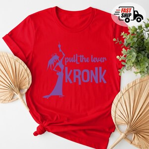 May include: A red t-shirt with the phrase "pull the lever KRONK" in purple. The design includes a silhouette of a character in a long dress. The shirt is folded at the bottom.