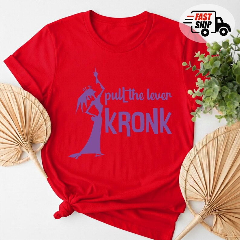 May include: A red t-shirt with the phrase "pull the lever KRONK" in purple. The design includes a silhouette of a character in a long dress. The shirt is folded at the bottom.