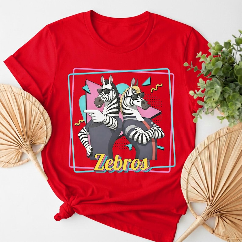 May include: Red t-shirt featuring a graphic of two zebras wearing sunglasses and casual clothing, with the word "Zebros" in yellow. The zebras are set against a retro-style background with a pink and blue design.