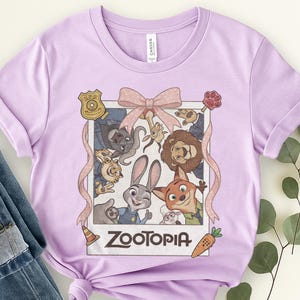 K&ouml;nnte beinhalten: Helllila T-Shirt mit einem Zootopia-Film-Motiv. Das Design zeigt ein Bild im Polaroid-Stil mit Charakteren wie Judy Hopps und Nick Wilde, eine rosa Schleife und das Wort "Zootopia".