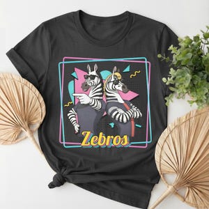 May include: Black t-shirt featuring a graphic of two zebras wearing sunglasses, with the word "Zebros" in yellow. The zebras are inside a pink and blue outlined rectangle with a retro design.