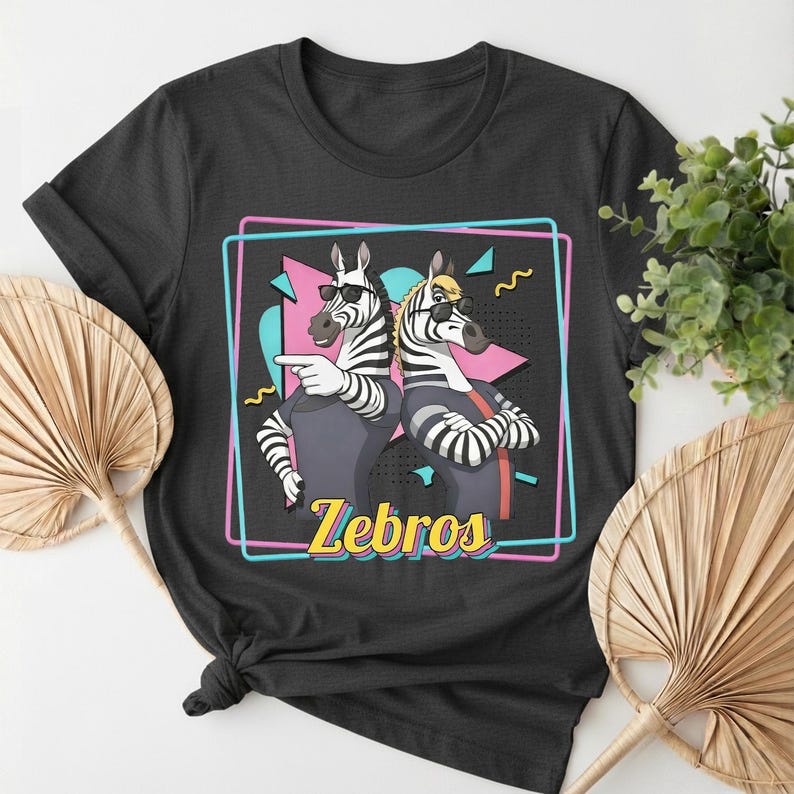May include: Black t-shirt featuring a graphic of two zebras wearing sunglasses, with the word "Zebros" in yellow. The zebras are inside a pink and blue outlined rectangle with a retro design.