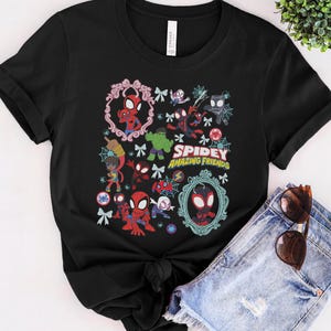 May include: Black t-shirt featuring a colorful graphic of Spidey and his Amazing Friends. The design includes various characters, decorative frames, and the text "Spidey Amazing Friends". Sunglasses and denim shorts are visible.