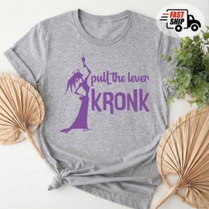 May include: A heather gray t-shirt featuring a purple graphic of a character and the text "pull the lever KRONK". The design includes a silhouette of a character in a dramatic pose. The shirt is made of a soft material.
