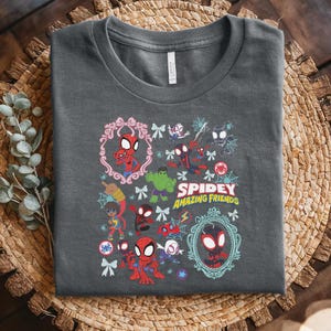 May include: Dark gray t-shirt featuring a colorful graphic of Spider-Man and friends. The design includes various characters, decorative frames, and the text "Spidey Amazing Friends". The shirt is folded on a woven surface.