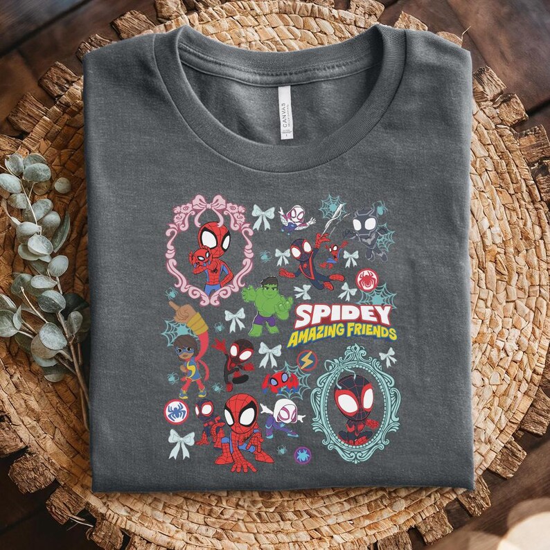 May include: Dark gray t-shirt featuring a colorful graphic of Spider-Man and friends. The design includes various characters, decorative frames, and the text "Spidey Amazing Friends". The shirt is folded on a woven surface.