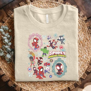 May include: Beige t-shirt featuring a colorful graphic of Spider-Man and other characters. The design includes the text "Spidey Amazing Friends" and various framed illustrations of the characters. The shirt is folded on a woven surface.