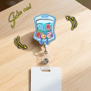 May include: A blue and white badge reel with a clear plastic ID holder. The badge reel features a whimsical underwater scene with a dolphin, octopus, seahorse, and other sea creatures. The text "Shake me!" is written in yellow.