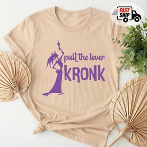 May include: Beige t-shirt with a purple graphic of a character and the text "pull the lever KRONK". The shirt has short sleeves and a crew neck. The design features a silhouette of a character in a long dress.