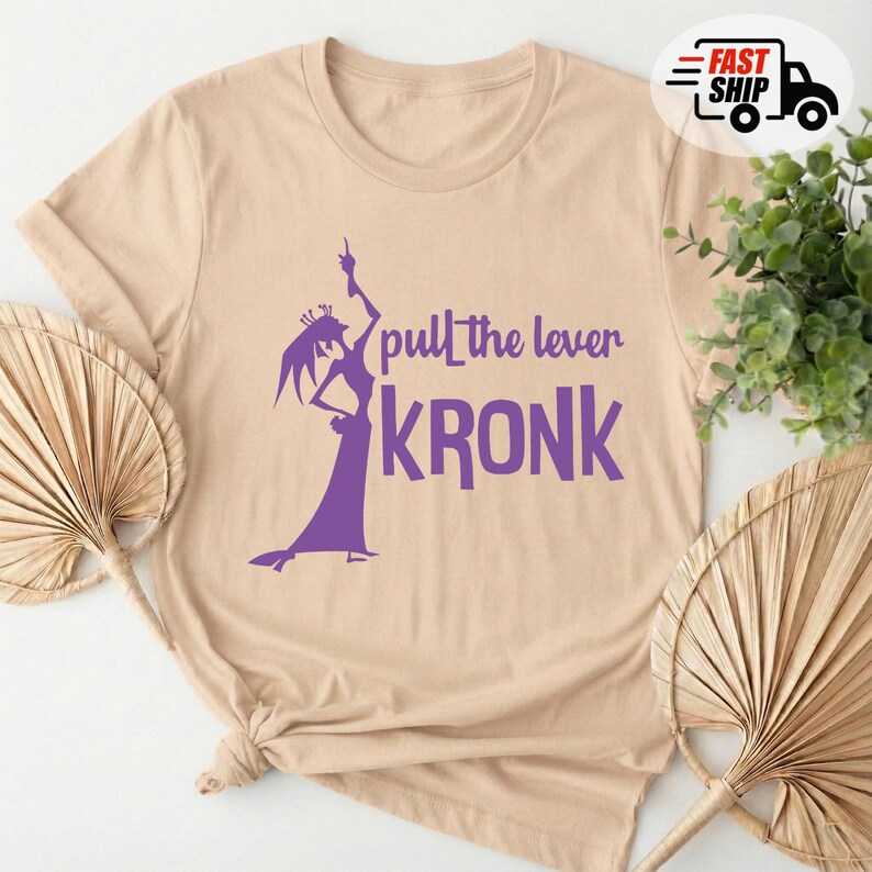 May include: Beige t-shirt with a purple graphic of a character and the text "pull the lever KRONK". The shirt has short sleeves and a crew neck. The design features a silhouette of a character in a long dress.