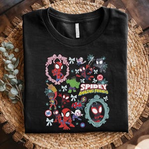 May include: Black t-shirt featuring a colorful graphic of Spider-Man and other characters. The design includes the text "Spidey and His Amazing Friends" with decorative frames, bows, and spiderwebs. The shirt is folded on a woven surface.