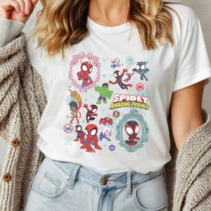 May include: White t-shirt featuring a colorful graphic of various Spider-Man characters and the text "Spidey Amazing Friends." The design includes cartoon illustrations of Spider-Man, Hulk, and other heroes in a playful style.