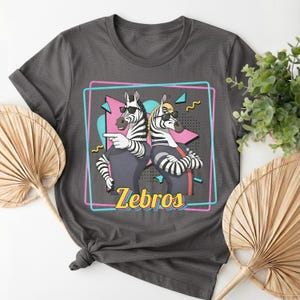 May include: Dark gray t-shirt featuring a graphic of two zebras wearing sunglasses, with the word "Zebros" in yellow. The zebras are inside a pink and blue rectangular frame, with a retro 80s design.
