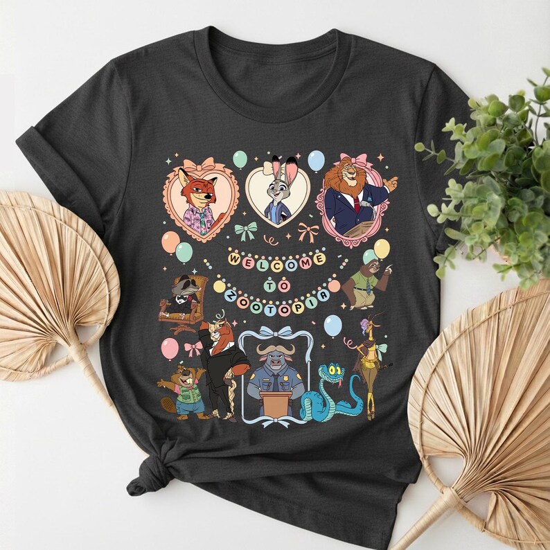 Puede incluir: Camiseta gris oscuro con un gr&aacute;fico colorido de personajes de Zootopia. El dise&ntilde;o incluye el texto "Welcome to Zootopia" y varios personajes de dibujos animados en marcos con forma de coraz&oacute;n. Globos y elementos decorativos rodean a los personajes.