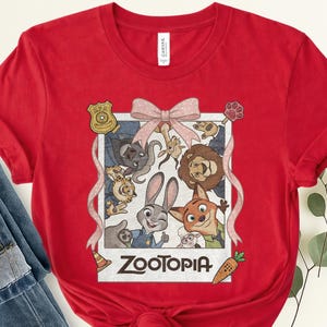K&ouml;nnte beinhalten: Rotes T-Shirt mit einem Grafikdruck von Charakteren aus dem Film "Zoomania". Das Design zeigt Judy Hopps, Nick Wilde und andere Charaktere in einem Polaroid-Rahmen, mit dem Filmtitel unten. Eine rosa Schleife und B&auml;nder schm&uuml;cken die Oberseite.