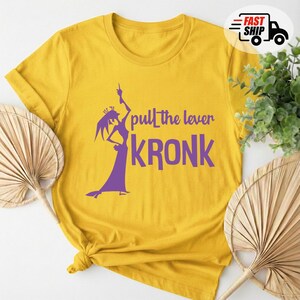 May include: Mustard yellow t-shirt with a purple graphic of a character and the text "pull the lever KRONK". The shirt is made of a soft material and has a crew neck.