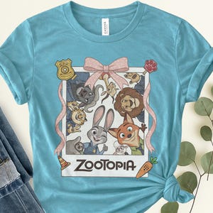 K&ouml;nnte beinhalten: Hellblaues T-Shirt mit einem gerahmten Bild von Charakteren aus dem Animationsfilm "Zoomania". Das Bild enth&auml;lt eine rosa Schleife, eine Polizeimarke und das Wort "Zootopia" in Wei&szlig;. Das Shirt ist aus weichem Material.