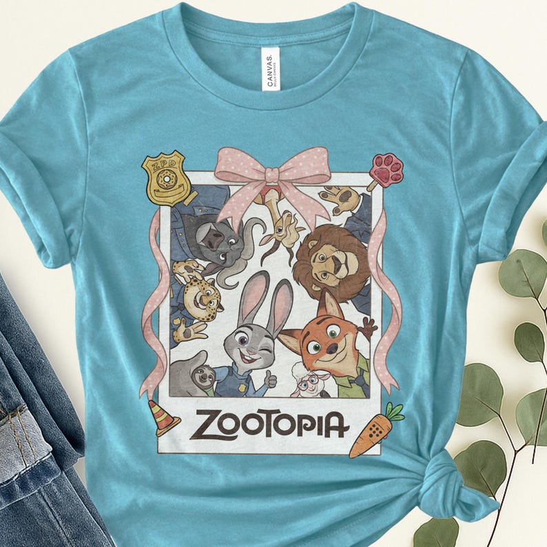 K&ouml;nnte beinhalten: Hellblaues T-Shirt mit einem gerahmten Bild von Charakteren aus dem Animationsfilm "Zoomania". Das Bild enth&auml;lt eine rosa Schleife, eine Polizeimarke und das Wort "Zootopia" in Wei&szlig;. Das Shirt ist aus weichem Material.