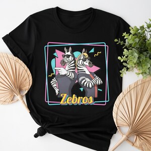 May include: Black t-shirt featuring a graphic of two zebras wearing sunglasses and casual attire, with the word "Zebros" in yellow. The zebras are set against a pink, blue, and black geometric background.