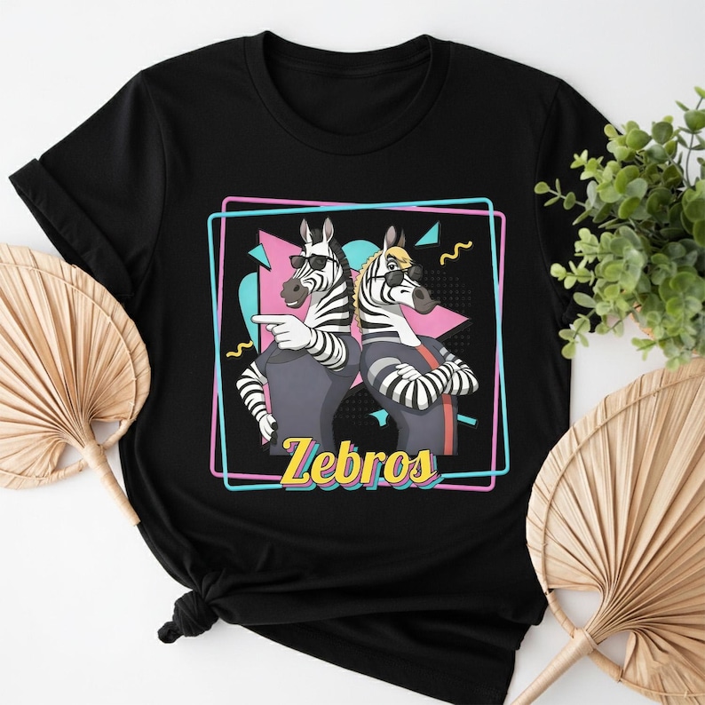 May include: Black t-shirt featuring a graphic of two zebras wearing sunglasses and casual attire, with the word "Zebros" in yellow. The zebras are set against a pink, blue, and black geometric background.