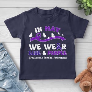 May include: Navy blue t-shirt with the text "IN MAY WE WEAR BLUE & PURPLE" and the hashtag "#Pediatric Stroke Awareness". The design includes a purple ribbon, butterflies, and text in white and purple.