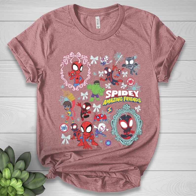 May include: A dusty rose t-shirt featuring a colorful graphic of various Spider-Man characters and the text "Spidey and His Amazing Friends." The design includes cartoon illustrations of Spider-Man, Hulk, and other heroes, framed by decorative elements.