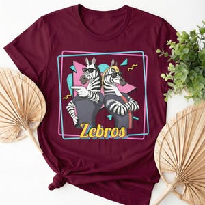 May include: Burgundy t-shirt featuring a cartoon illustration of two zebras wearing sunglasses and casual attire. The zebras are inside a pink and blue outlined box with the word "Zebros" in yellow. The shirt has rolled-up sleeves.