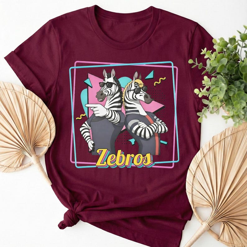 May include: Burgundy t-shirt featuring a cartoon illustration of two zebras wearing sunglasses and casual attire. The zebras are inside a pink and blue outlined box with the word "Zebros" in yellow. The shirt has rolled-up sleeves.
