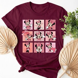 May include: A burgundy t-shirt displaying a grid of Minnie Mouse illustrations. Each square features Minnie in different poses, wearing a pink polka-dot dress and bow. The design is a cheerful and colourful tribute to the iconic character.