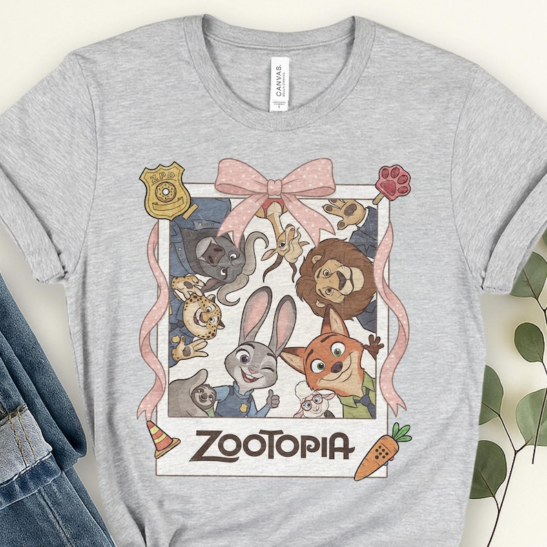 K&ouml;nnte beinhalten: Hellgraues T-Shirt mit einer farbenfrohen Grafik aus dem Film Zootopia. Die Grafik zeigt verschiedene Tierfiguren, eine rosa Schleife und das Wort "Zootopia" in fetter Schrift. Eine Karotte und ein Verkehrskegel sind ebenfalls im Design enthalten.