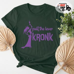 May include: Dark green t-shirt with a purple graphic of a character and the text "pull the lever KRONK". The shirt is made of a soft material and has a classic crew neck design. A fun and unique graphic tee.