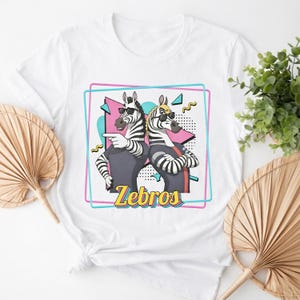 May include: White t-shirt featuring a graphic of two zebras wearing sunglasses, with the word "Zebros" in yellow. The zebras are set against a pink and blue geometric design.