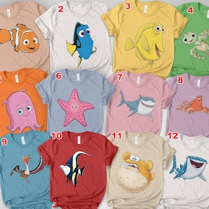 May include: A collection of twelve short-sleeved t-shirts in various colors, each featuring a cartoon sea creature graphic. Designs include a clownfish, a blue tang, a yellow tang, a sea turtle, an octopus, a starfish, a shark, an octopus, a pelican, a butterfly fish, a pufferfish, and a whale shark.