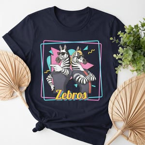 May include: Navy blue t-shirt featuring a graphic of two zebras wearing sunglasses, with the word "Zebros" in yellow. The zebras are set against a retro-style background with pink, blue, and black shapes.
