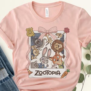 K&ouml;nnte beinhalten: Hellrosa T-Shirt mit einem Aufdruck von Charakteren aus dem Film Zootopia. Die Grafik ist mit einem rosa Band umrahmt und enth&auml;lt das Wort "Zootopia". Das T-Shirt besteht aus einem weichen Material.