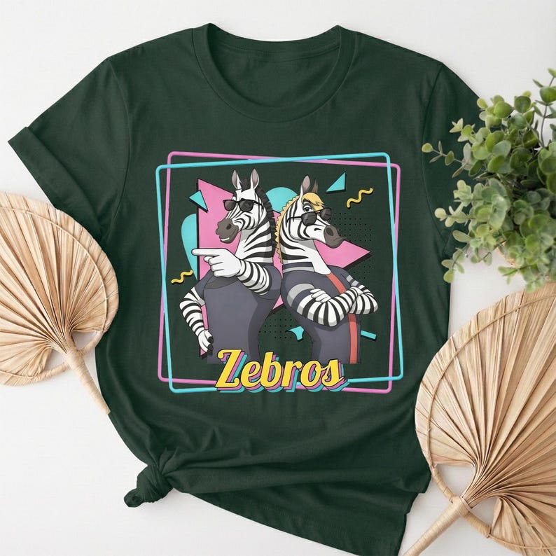 May include: Dark green t-shirt featuring a graphic of two zebras wearing sunglasses, with the word "Zebros" in yellow and orange. The zebras are inside a pink and blue outlined rectangle. The shirt has short sleeves.