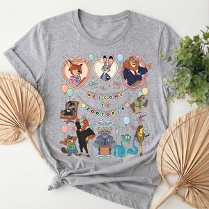 Puede incluir: Una camiseta gris con un dise&ntilde;o colorido con personajes de la pel&iacute;cula Zootopia. El dise&ntilde;o incluye el texto "WELCOME TO ZOOTOPIA" y varios personajes de animales en marcos con forma de coraz&oacute;n y otros elementos decorativos. La camiseta es de material suave.
