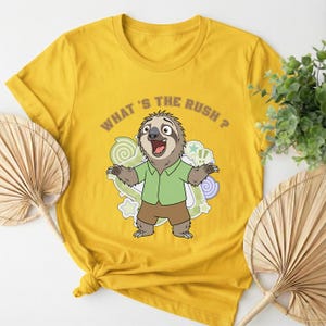 May include: Mustard yellow t-shirt featuring a cartoon sloth with the text "WHAT'S THE RUSH?". The sloth is wearing a green shirt and brown shorts, with a surprised expression and outstretched arms. The design includes swirls and stars.