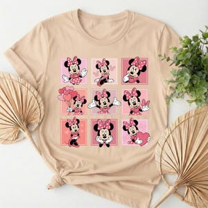 May include: A beige t-shirt with a grid of nine Minnie Mouse illustrations. Each Minnie Mouse is depicted in a pink dress and bow. The background squares alternate between pink and light pink.